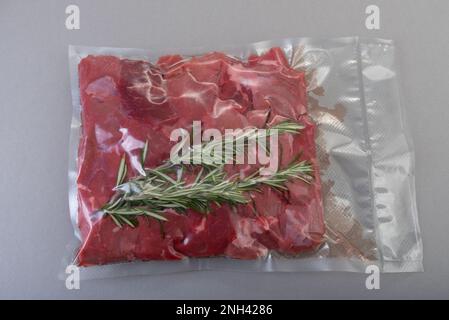 Viande de bœuf crue coupée en dés pour ragoût et branche de romarin dans un emballage scellé sous vide pour la cuisson sous vide sur fond gris Banque D'Images