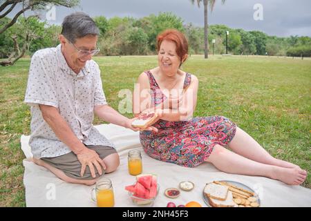 Un heureux couple latin senior ayant un petit déjeuner pique-nique avec du pain aux grains entiers et des biscuits secs, jus d'orange et fruits variés. Banque D'Images