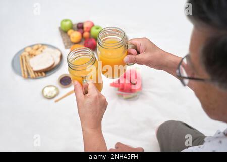 Un couple plus âgé toaster avec du jus d'orange frais tout en dégustant un pique-nique sain avec du pain aux grains entiers, des biscuits secs et des fruits variés. Banque D'Images