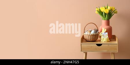 Panier avec œufs de Pâques, lapin et tulipes jaunes sur table en bois près du mur beige. Bannière pour le design Banque D'Images