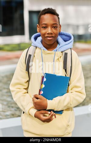 Jeune garçon afro-américain souriant avec des livres d'imitation et un sac à dos debout devant l'appareil photo dans un environnement urbain Banque D'Images