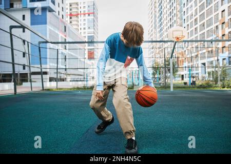 Élève actif au secondaire dans le pantalon et le pull-over jouant au basket-ball sur le terrain de sport après les cours ou à loisir contre les maisons Banque D'Images