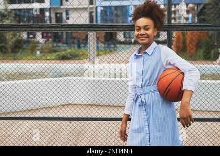 Bonne et réussie écolière en robe décontractée tenant le ballon tout en se tenant contre le filet de basket-ball sur le terrain de jeu et en regardant l'appareil photo Banque D'Images