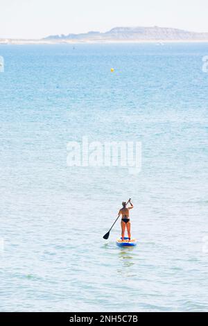 BOURNEMOUTH, Royaume-Uni - 08 juillet 2022. Femme d'âge mûr ou moyen avec des cheveux gris paddle-board au large de la côte Dorset. Avec l'île de Wight à l'horizon Banque D'Images