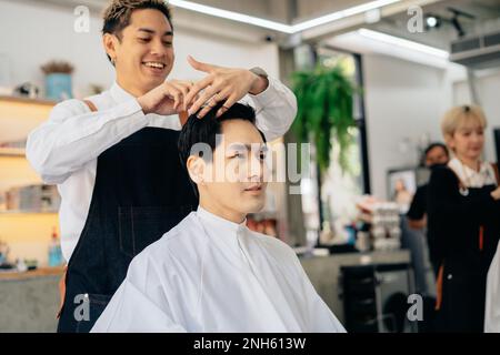 Joyeux coiffeur de jeune asiatique souriant portant un tablier dans moderne salon tenant la brosse et les ciseaux de couper les cheveux de client Banque D'Images