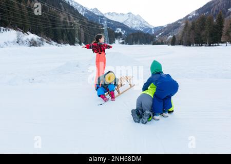 groupe d'enfants en vêtements d'hiver lumineux jouant avec un traîneau en bois sur la vallée de la neige sur fond de forêt et de montagne Banque D'Images