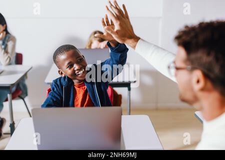 Joyeux gamin noir qui fait cinq hauts avec son professeur dans une salle de classe informatique, célébrant un moment d'enseignement et d'apprentissage réussi. Jeune stude Banque D'Images