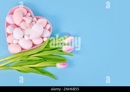 Décoration de Pâques avec œufs peints dans une assiette en forme de coeur et fleurs de tulipe roses sur fond bleu avec espace de copie Banque D'Images