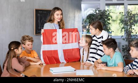 Le professeur d'école parle aux élèves du Danemark et détient un drapeau du Danemark entre les mains. Banque D'Images