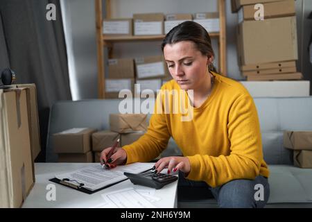 Femme entrepreneur sérieux utilisant la calculatrice avec stylo, calcul des dépenses financières et budget à la maison bureau, marché en ligne emballage boîte livraison, sta Banque D'Images
