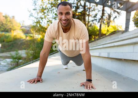 Homme hispanique souriant et gai faisant de l'exercice en plein air dans le stade, homme s'étirant et se récupérant avant de courir dans le parc d'été. Banque D'Images