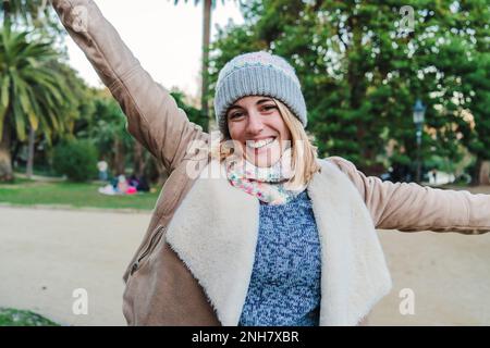 Vue de face d'une jeune femme blonde de race blanche souriante aux dents blanches parfaites, portant un bonnet beanie et regardant excitée de la caméra dans un parc à l'extérieur. Photo de haute qualité Banque D'Images