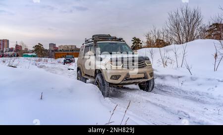 Les véhicules tout-terrain russes UAZ Patriot, blancs et sombres, roulent rapidement sur une route enneigée dans un champ en hiver, accrochant à une tempête de neige avec une roue. Banque D'Images