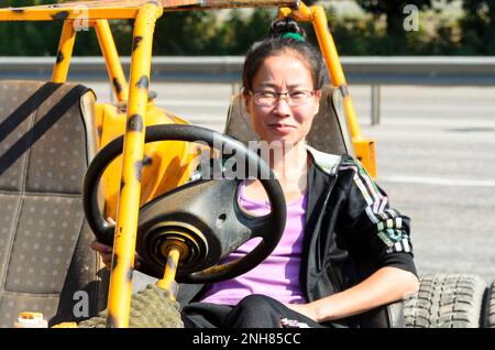 Fille en costume 'Adidas' amusement est assis derrière le volant d'une petite voiture buggy à côté de la publicité de la station de service Banque D'Images