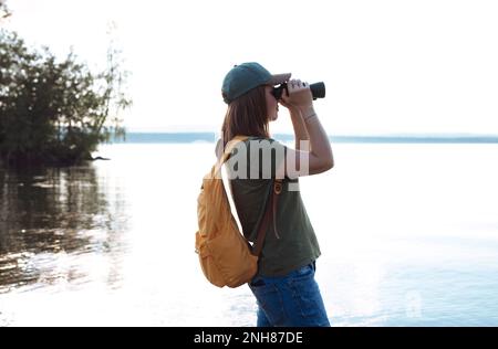 Jeune femme avec sac à dos jaune regardant à travers des jumelles aux oiseaux sur le lac ou la rivière observation des oiseaux, zoologie, écologie recherche dans la nature, observation o Banque D'Images