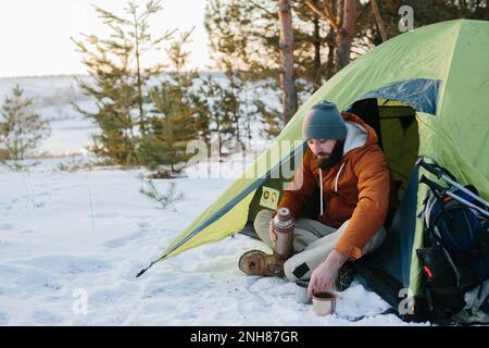 Un jeune homme barbu repose dans les montagnes d'hiver près d'une tente. Un homme-voyageur avec une barbe dans une casquette et une veste chaude se réchauffe en buvant du thé chaud ou Banque D'Images