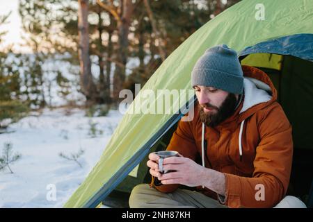 Un jeune homme barbu repose dans les montagnes d'hiver près d'une tente. Un homme-voyageur avec une barbe dans une casquette et une veste chaude se réchauffe en buvant du thé chaud ou Banque D'Images