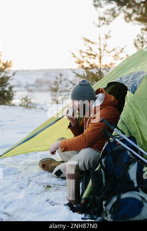 Un jeune homme barbu repose dans les montagnes d'hiver près d'une tente. Un homme-voyageur avec une barbe dans une casquette et une veste chaude se réchauffe en buvant du thé chaud ou Banque D'Images
