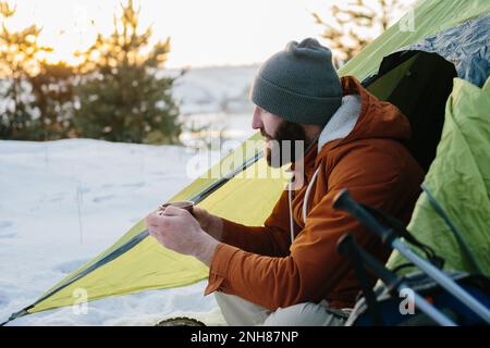 Un jeune homme barbu repose dans les montagnes d'hiver près d'une tente. Un homme-voyageur avec une barbe dans une casquette et une veste chaude se réchauffe en buvant du thé chaud ou Banque D'Images