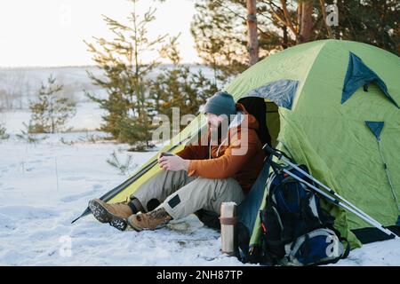 Un jeune homme barbu repose dans les montagnes d'hiver près d'une tente. Un homme-voyageur avec une barbe dans une casquette et une veste chaude se réchauffe en buvant du thé chaud ou Banque D'Images