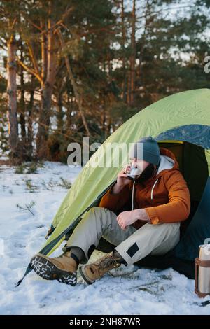 Un jeune homme barbu repose dans les montagnes d'hiver près d'une tente. Un homme-voyageur avec une barbe dans une casquette et une veste chaude se réchauffe en buvant du thé chaud ou Banque D'Images
