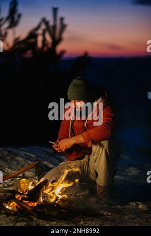 Un randonneur vérifie son téléphone tout en se réchauffant par un feu de camp la nuit dans les montagnes. Banque D'Images