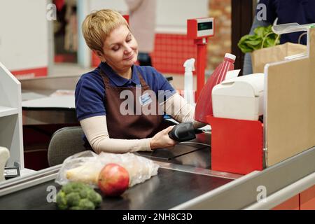 Blonde jolie femme caissière en uniforme balayant le prix des produits dans une bouteille rouge tout en étant assise au comptoir des caisses et en travaillant Banque D'Images