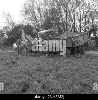 1950s, historique, un camion avec une charge pleine de gravats et de terre avec ses roues bloquées sur le côté d'une piste de ferme boueuse, Angleterre, Royaume-Uni. Plusieurs ouvriers agricoles avec des bêches essayant de creuser le véhicule. Un creuseur est devant le chariot. Banque D'Images