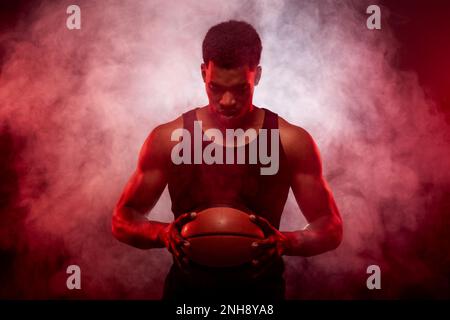 Côté joueur de basket-ball éclairé de couleur rouge tenant une balle sur fond de fumée. Sérieux homme afro-américain concentré. Banque D'Images