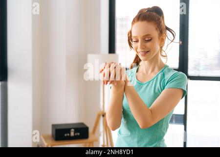 Portrait de taille moyenne de la jeune femme souriante faisant l'effort d'étirement poignet avant l'entraînement de fitness au bureau à la maison près de la fenêtre. Banque D'Images