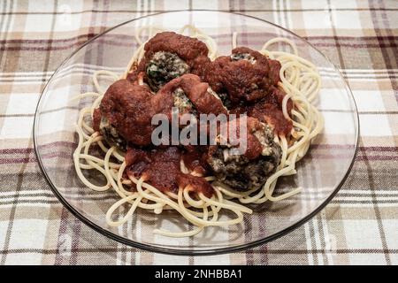 Boulettes de viande de venaison sur de longues nouilles spaghetti recouvertes d'une sauce spaghetti sur une plaque de verre transparent sur une nappe de tissu écossais brun. Banque D'Images