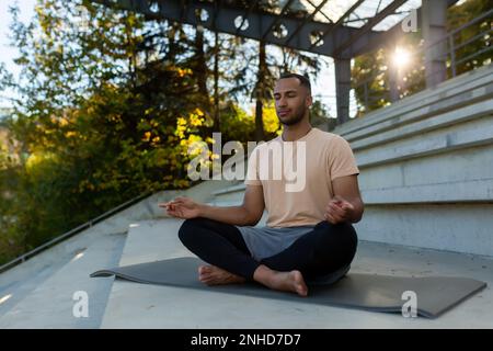 Un jeune Africain américain est assis sur un tapis dans un stade en position lotus et fait du yoga. Il médite, ferme les yeux, détendu, calme. Banque D'Images