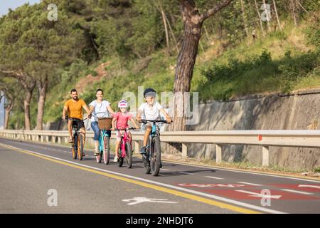 Deux enfants, avec un casque sur leur tête, et parents souriants à vélo sur une piste cyclable familiale le long d'une côte de mer, vue de face. Banque D'Images