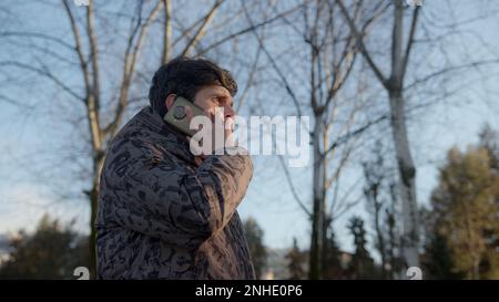 Un jeune homme confiant et professionnel dans une élégante veste de sport noire engagé dans un appel téléphonique sérieux en plein air, démontrant le dévouement et le reliabilit Banque D'Images