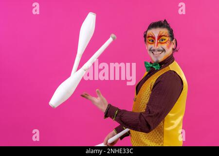 Clown avec maquillage blanc pour le visage sur fond rose, clubs de jonglage souriants et regardant l'appareil photo Banque D'Images