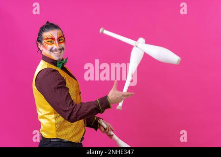 Clown avec maquillage blanc pour le visage sur fond rose, clubs de jonglage souriants et regardant l'appareil photo Banque D'Images