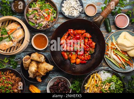 Ensemble de plats chinois variés sur la table : poulet aigre-doux dans un wok, dim sum dans un cuiseur vapeur en bambou, rouleaux de printemps, nouilles, salade, riz, vapeur Banque D'Images