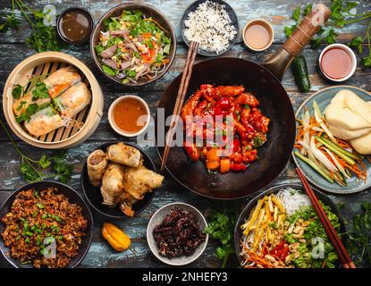 Ensemble de plats chinois variés sur la table : poulet aigre-doux dans un wok, dim sum dans un cuiseur vapeur en bambou, rouleaux de printemps, nouilles, salade, riz, vapeur Banque D'Images