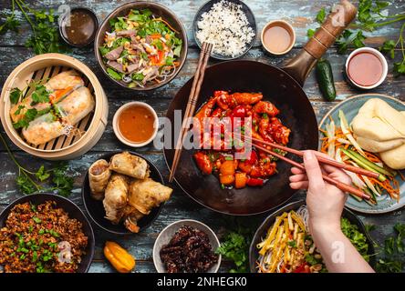 Ensemble de plats chinois variés sur la table : poulet aigre-doux dans un wok, dim sum dans un cuiseur vapeur en bambou, rouleaux de printemps, nouilles, salade, riz, vapeur Banque D'Images