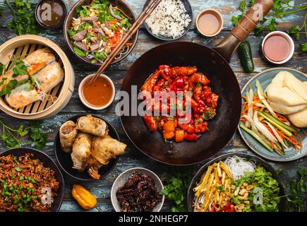 Ensemble de plats chinois variés sur la table : poulet aigre-doux dans un wok, dim sum dans un cuiseur vapeur en bambou, rouleaux de printemps, nouilles, salade, riz, vapeur Banque D'Images