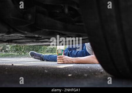 Un mécanicien au sol, on peut voir sa jambe et son bras tenir un produit. La photo a été prise sous la voiture qui permet de voir une roue. Banque D'Images