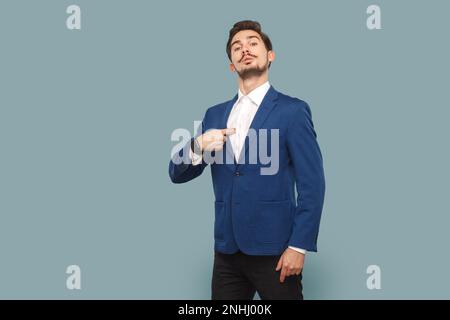 Portrait d'un homme fier et sûr de lui avec une moustache qui pointe vers lui, regardant l'appareil photo avec fierté, portant une chemise et une veste blanches. Prise de vue en studio isolée sur fond bleu clair. Banque D'Images