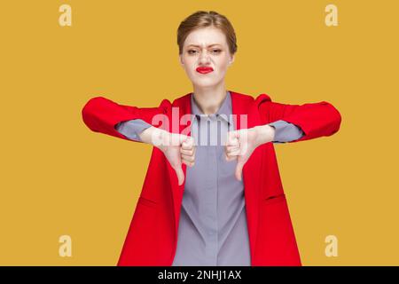 Portrait de triste bouleversé jeune femme adulte avec des lèvres rouges debout et montrant les pouces vers le bas, n'aime pas le service, démontrer désapprouver, portant la veste rouge. Studio d'intérieur isolé sur fond jaune. Banque D'Images