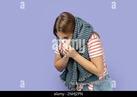 Portrait de femme blonde allergique portant un T-shirt rayé et enveloppé dans un foulard souffrant de nez liquide, ayant la température élevée, éternuant, attrapant le froid. Studio d'intérieur isolé sur fond violet. Banque D'Images