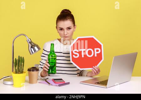 Portrait d'une femme impatiente montrant une bouteille de bière alcoolisée et un panneau d'arrêt, assise sur le lieu de travail avec un ordinateur portable. Studio d'intérieur tourné isolé sur fond jaune. Banque D'Images