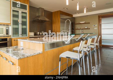 Armoires en bois et en verre et îlot avec comptoir en granit nuancé gris et chaises hautes en cuir blanc dans la cuisine avec sol carrelé en marbre gris. Banque D'Images