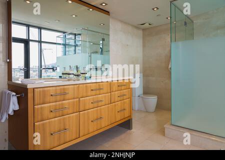 Meuble-lavabo en bois exotique et cabine de douche en verre dans la salle de bains attenante avec sol carrelé en marbre à l'intérieur d'un luxueux appartement de grand standing moderne de plusieurs étages. Banque D'Images