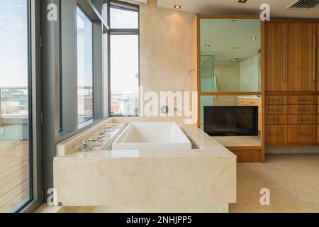 Baignoire blanche avec boîtier en marbre et miroir avec télévision grand écran intégrée dans la salle de bains attenante avec sol carrelé en marbre dans un luxueux appartement moderne. Banque D'Images