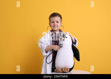 Mignon petit garçon en uniforme de pédiatre jouant avec stéthoscope et pingouin jouet sur fond jaune Banque D'Images