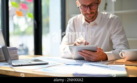 Travailleur de bureau ou analyste financier spécialisé dans le Caucase, intelligent et concentré, utilisant une tablette numérique, travaillant sur un rapport de données financières à son bureau. Banque D'Images